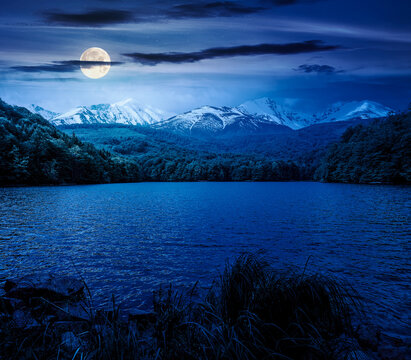mountain lake in springtime at night. composite landscape of lake and ridge in full moon light. calm water, green forest and snow capped peaks. backdrop for fiction art or mystery concepts