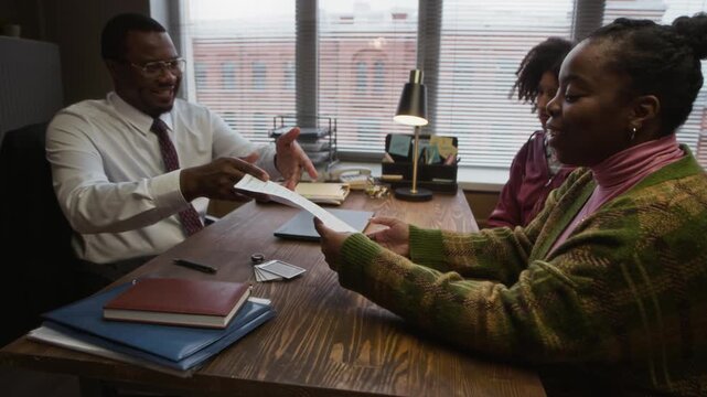 Handheld shot of male principal giving test with excellent result to mother of teenage student during appointment in modern school office