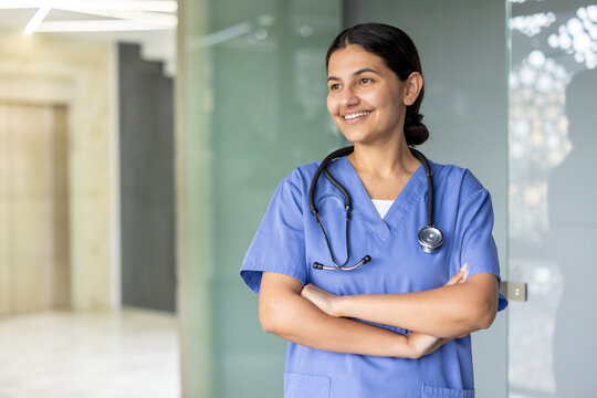 Female healthcare professional standing with arms crossed, smiling while looking away, wearing blue scrubs and a stethoscope in a hospital or clinic setting, representing dedication and care