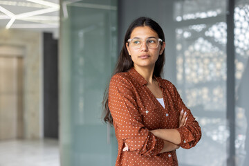 Young professional indian woman wearing glasses and a polka dot shirt, standing with arms crossed...