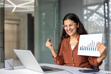Happy businesswoman discussing positive chart data during an online video conference, gesturing excitedly while presenting good news about business growth and achievement from the modern office