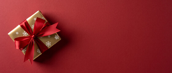 A gold wrapped gift box with a red bow rests on a deep red banner background, conveying festive...