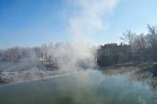 A dramatic winter scene of the Kupa River dam in Ozalj, where the historic Munjara hydroelectric power plant is visible on the right, surrounded by heavy fog and frost-covered tree