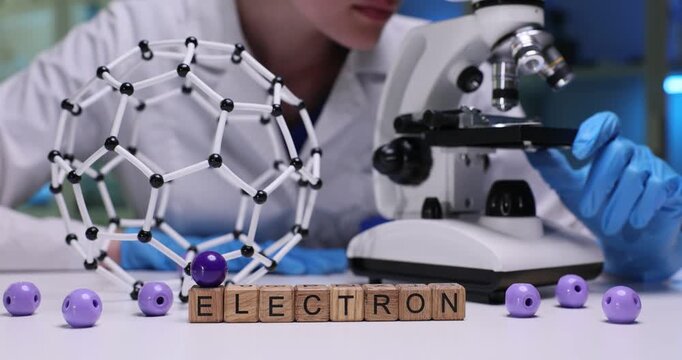 Woman lab technician studies prepared sample through microscope lens. Word Electron formed from wooden cubes positioned neatly on work surface