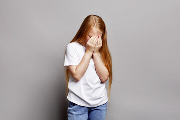 Young girl in studio covering face with hands, looking upset and vulnerable isolated on gray background. Expressive portrait stressed child, teenage emotional struggles and adolescent difficulties