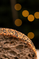 A close-up of sliced artisan sourdough bread with a porous, airy crumb and a crunchy crust coated in black and golden sesame seeds framed by a dark background illuminated with warm bokeh lights.