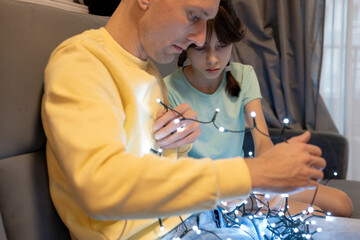 Father and daughter untangling glowing Christmas lights while decorating at home, warm festive family moment