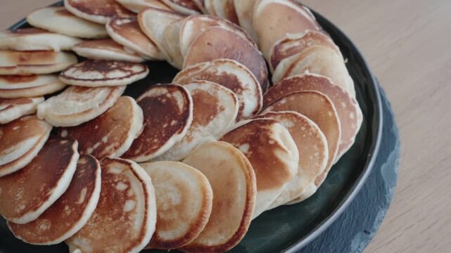 A plate full of small homemade pancakes arranged in a circular pattern on a table. Soft natural light creates a warm and cozy breakfast atmosphere.