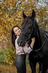 Obraz premium A young Caucasian woman with long dark hair hugs a large black horse in a colorful autumn setting with trees and fallen leaves.