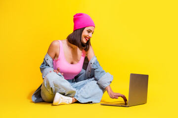 Young female model in pink beanie denim streetwear sits with laptop on bright yellow background smiling and enjoying a fashion inspired moment