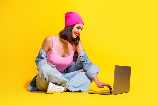 Young female model in pink beanie denim streetwear sits with laptop on bright yellow background smiling and enjoying a fashion inspired moment