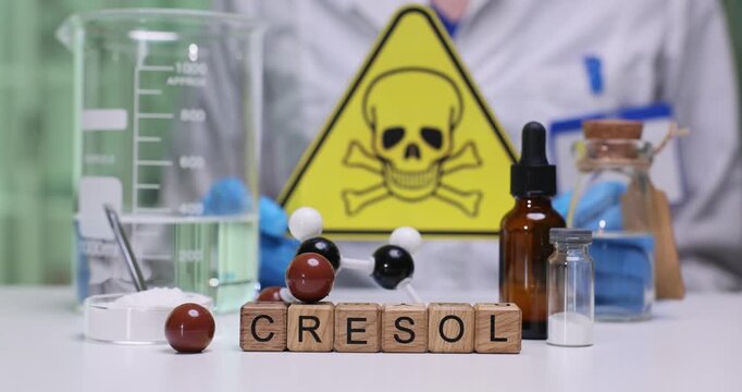 Chemist shows triangular caution sign to indicate hazardous chemicals. Wooden cubes on desk form letters of word Cresol in precise row among glassware