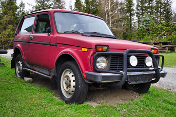 Wytrzymały, stary czerwony SUV stoi zaparkowany na trawie obok lasu. Wyróżnia się masywnym zderzakiem i okrągłymi reflektorami, przywodząc na myśl eksplorację przyrody, nostalgię i surową przygodę. © Pol Badger