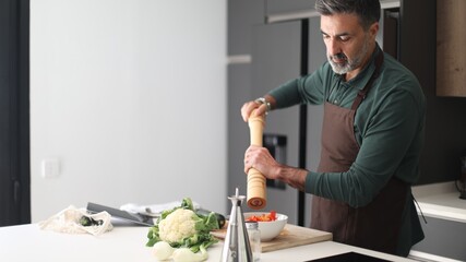 Man seasoning fresh vegetables with pepper grinder in kitchen