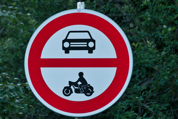 Znak drogowy zakazujący wjazdu samochodom i motocyklom na tle zieleni. To zdjęcie stockowe oddaje bezpieczeństwo na drodze, przepisy ruchu drogowego i kontekst środowiskowy. © Pol Badger