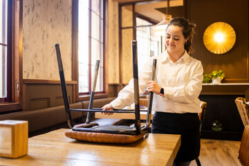 A young woman between 20 and 30 years old with Down syndrome works as a waitress in a modern cafe.The girl is collecting the chairs to end her shift.Concept of trisomy 21.