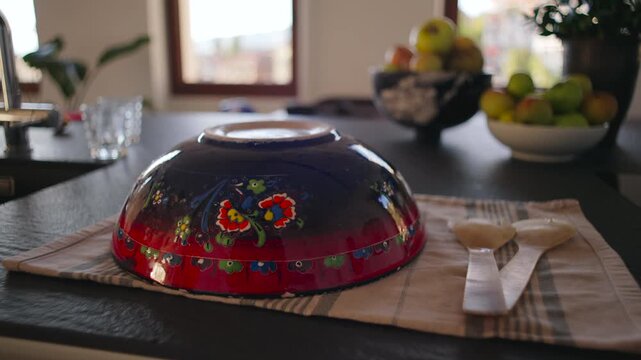 Decorated ceramic bowl resting upside down on cloth beside wooden spoons on kitchen counter with soft daylight creating warm domestic moment of quiet home preparation space