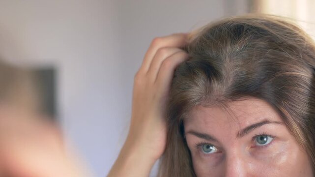 Closeup brunette woman checking roots in mirror, parting hair with focused motion, exploring home coloring options. Woman examining gray hairs in mirror, parting strands to study gray hairs 