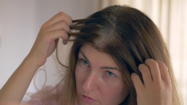 Woman checking gray hair in mirror, separating strands to inspect gray hairs and early root change. Concept video of young woman getting old. Natural aging signs and beauty concern useful for haircare