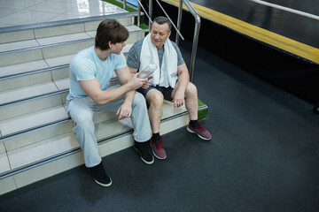 Adult man sitting in gym talking with younger trainer, resting after workout and receiving guidance for improving physical fitness. Senior Man Working Out in Gym