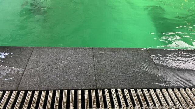 Vibrant green swimming pool water gently overflows a dark grey tiled edge into a drainage grate creating small ripples and a serene atmosphere