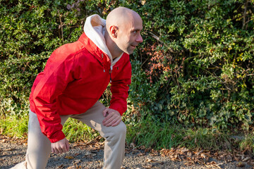 young man in red jacket legs and hamstrings, crouching on park path, performing pre run dynamic stretching, solo training in cold air, discipline and small daily victories, surrounded by green bushes
