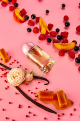 A perfume bottle sits among slices of peaches, berries, and flowers on a pink surface. The scene includes various fruits, adding a playful touch to the layout.