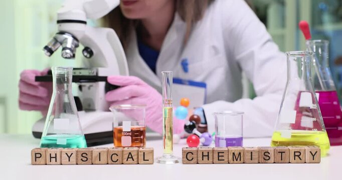 Miniature wooden cubes forming phrase Physical Chemistry put on table with glassware. Woman lab worker carefully examines specimen under microscope