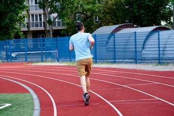 Obraz premium Man in casual attire jogging on a red track field surrounded by a city environment. Highlights wellness, activity, and urban lifestyle, combining fitness with an outdoor setting in a casual setting.