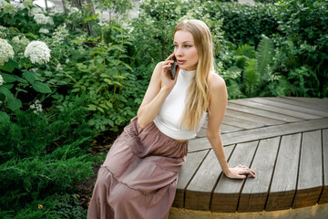 A beautiful blonde woman is talking on the phone in a city park. Communication, business relations.