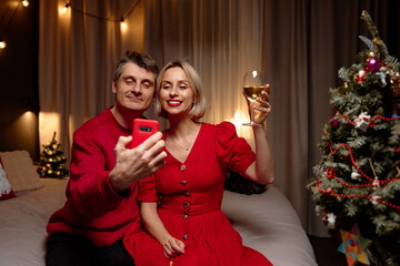 A smiling couple sits on a bed in a cozy living space, capturing a selfie with a glass of wine while celebrating Christmas beside a decorated tree. Warm lights create a festive mood.