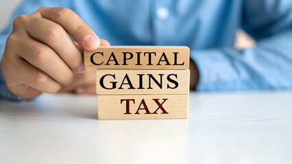 A man is holding wooden blocks with the words capital gains tax written on them on a white table