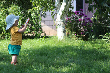 Child standing on green grass in a garden, wearing a white sun hat, yellow shirt, and green shorts, looking at a smartphone. Bright natural light and colorful flowers in the background create a cheerf