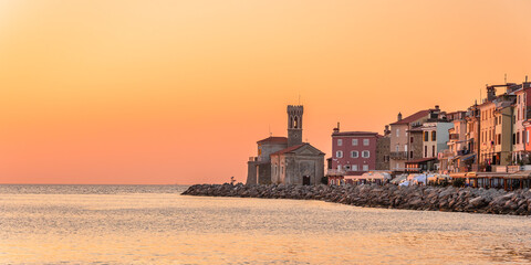 Obraz premium The coastal town of Piran in Slovenia is illuminated by the rays of the setting sun over the sea. The coastal landscape is visible from the shore, with an orange sky.