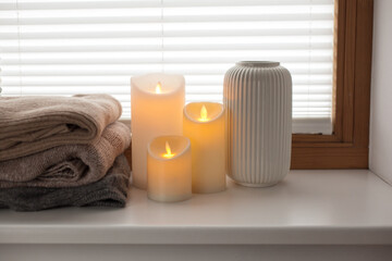 Pile of warm knitted sweaters, led battery candles and vase over white background with soft window light.