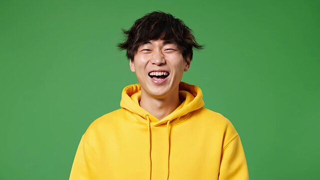 Young man in yellow hoodie laughing shyly looking down with amusement on a green screen Cheerful male giggling happily perfect for chroma key projects requiring