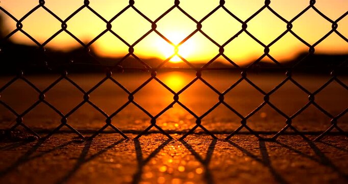 Golden sunset shines brightly through a chainlink fence, casting long shadows on the ground in this outdoor scene, evoking serenity and hope