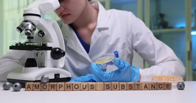 Chemistry laboratory worker studies tiny sample under high-precision microscope. Wooden blocks on table clearly form phrase Amorphous Substance
