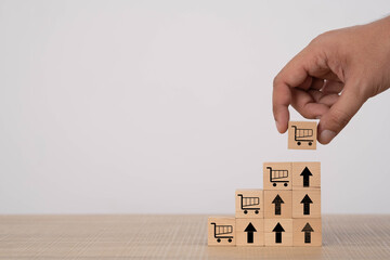 A hand arranging wooden blocks with shopping cart icons and upward arrows, symbolizing sales growth, e-commerce success, business development, increasing demand, and strategic online market expansion.