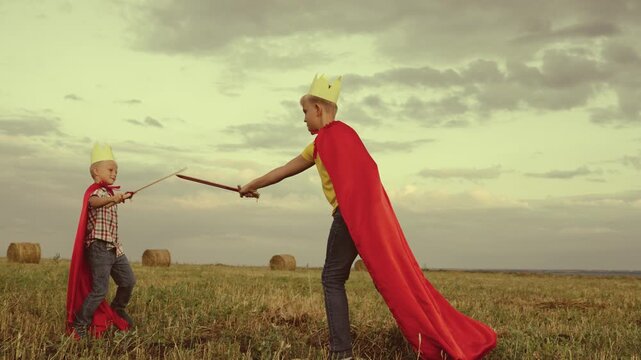 children superheroes play with swords field kings knights. dream childhood. little kid and his brother fearless defenders. teamwork child boys sons. dream adventure red capes at sunset. summer games