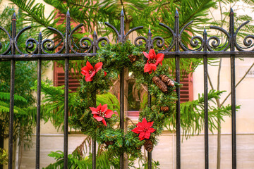 A festive Christmas wreath, decorated with red flowers and pinecones, hangs on a black wrought-iron fence against a background of Mediterranean tropical plants.
