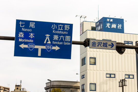 Kanazawa, Japan. Large blue road sign showing directions in Japanese and Latin script to Kenroku-en Garden, Morimoto, Nanao, Kodatsuno, and Kanazawa Castle on a cloudy day