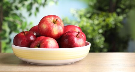 Ripe red apples in bowl on wooden table against blurred green background, closeup. Space for text