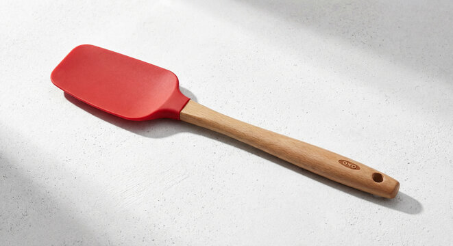 Modern red silicone kitchen spatula with a natural wooden handle, featuring the OXO brand, lying diagonally on a bright white textured surface with subtle shadows.