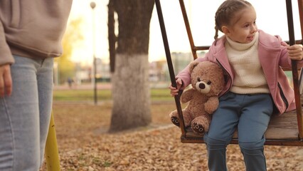 a little child sways with a teddy bear in an autumn park, a happy kid dreams of flying with his favorite toy, an active family vacation of mother and daughter on walk in city, childhood friend of girl