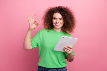 Young woman with curly hair in a green tshirt holds a tablet and makes an ok gesture against pink background for lifestyle fashion advertising and promotion