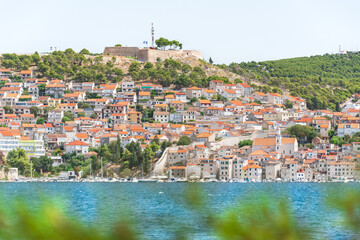 The coastal town of Sibenik in Croatia sits on the seafront, with hilltop houses and the Barone Fortress. A view from the other side on a sunny summer day.