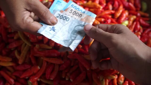 Close-up of a cash transaction using Indonesian rupiah at a traditional market, with hands exchanging money above a pile of fresh chili peppers. Perfect for themes of trade, economy, finance