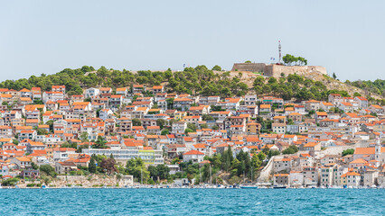 The coastal town of Sibenik in Croatia sits on the seafront, with hilltop houses and the Barone Fortress. A view from the other side on a sunny summer day.