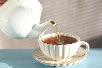 Woman pouring hot tea into cup from teapot at light blue wooden table, closeup